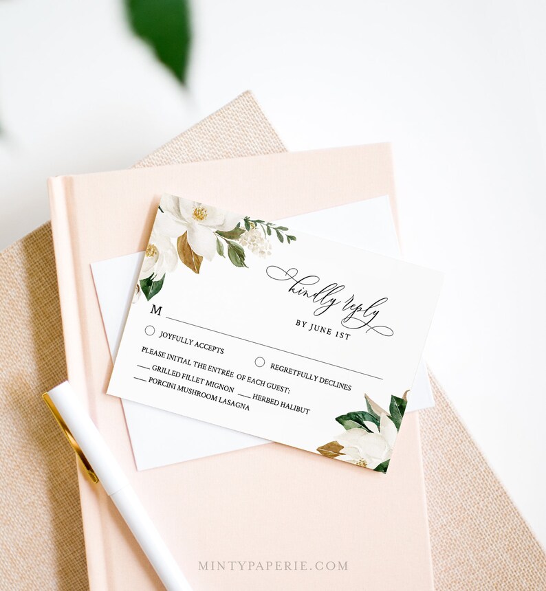 Puede incluir: Una tarjeta RSVP blanca con un dise&ntilde;o floral con flores de magnolia blancas y hojas verdes. La tarjeta tiene un encabezado negro que dice "Kindly Reply by June 1st". La tarjeta tiene dos opciones para que los invitados elijan: "Joyfully Accepts" y "Regretfully Declines".