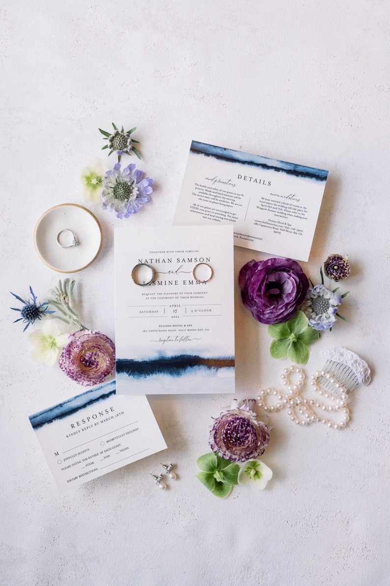 May include: Wedding invitation suite with blue watercolor accents. The invitation features the names "Nathan Samson and Jasmine Emma" with the date "April 17" and the time "5 o'clock". The invitation is surrounded by flowers and a pearl necklace.