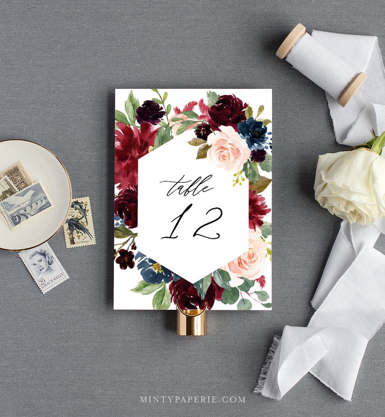May include: A white wedding table number card with a burgundy, blush, and navy floral design. The card features a hexagonal frame with the text "table 12" in black calligraphy.