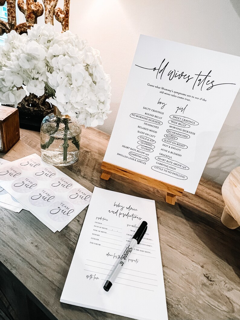 May include: A white sign with black text that reads "Old Wives Tales" and lists common pregnancy symptoms for both boys and girls. The sign is on a wooden easel and there are several stickers that say "Team Girl" on a table.
