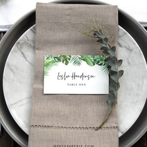May include: A white place card with a green tropical leaf border. The text on the card reads "Leslie Henderson, Table One".