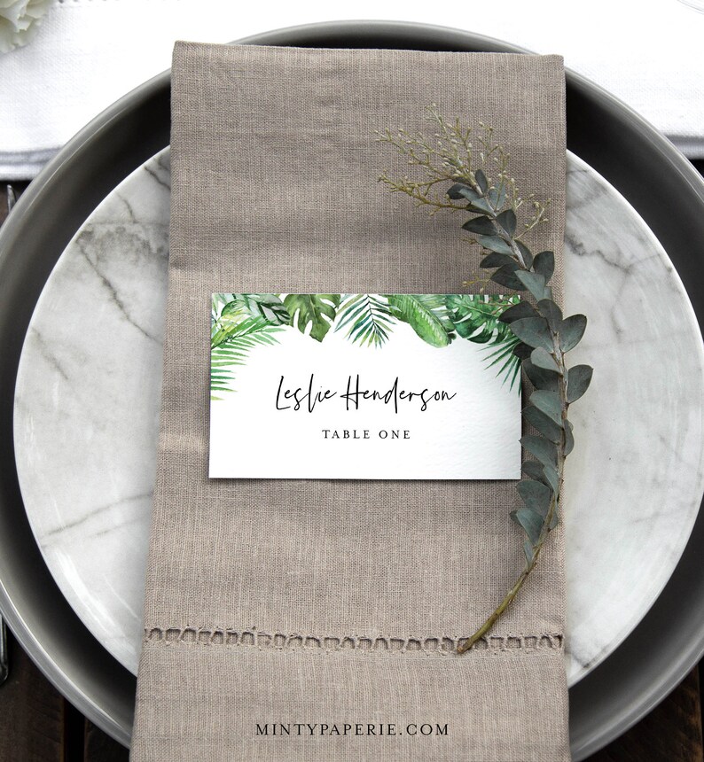 May include: A white place card with a green tropical leaf border. The text on the card reads "Leslie Henderson, Table One".