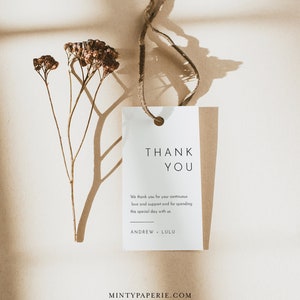May include: A white thank you tag with a brown string attached. The tag has the text "THANK YOU" in black lettering. The text below reads "We thank you for your continuous love and support and for spending this special day with us. ANDREW LULU". The tag is on a brown surface with a dried flower stem.