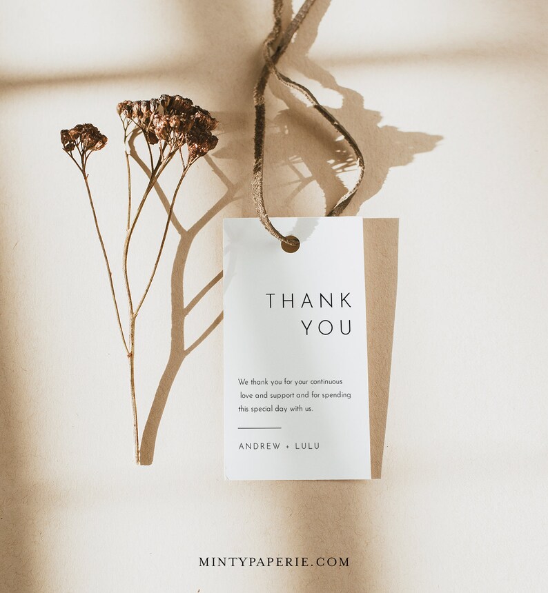 May include: A white thank you tag with a brown string attached. The tag has the text "THANK YOU" in black lettering. The text below reads "We thank you for your continuous love and support and for spending this special day with us. ANDREW LULU". The tag is on a brown surface with a dried flower stem.