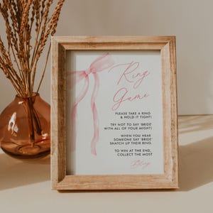 May include: A framed sign with the text "Ring Game" in pink script. The sign includes instructions and a watercolor-style pink bow. The frame is made of light-colored wood, and a vase with dried flowers is in the background.