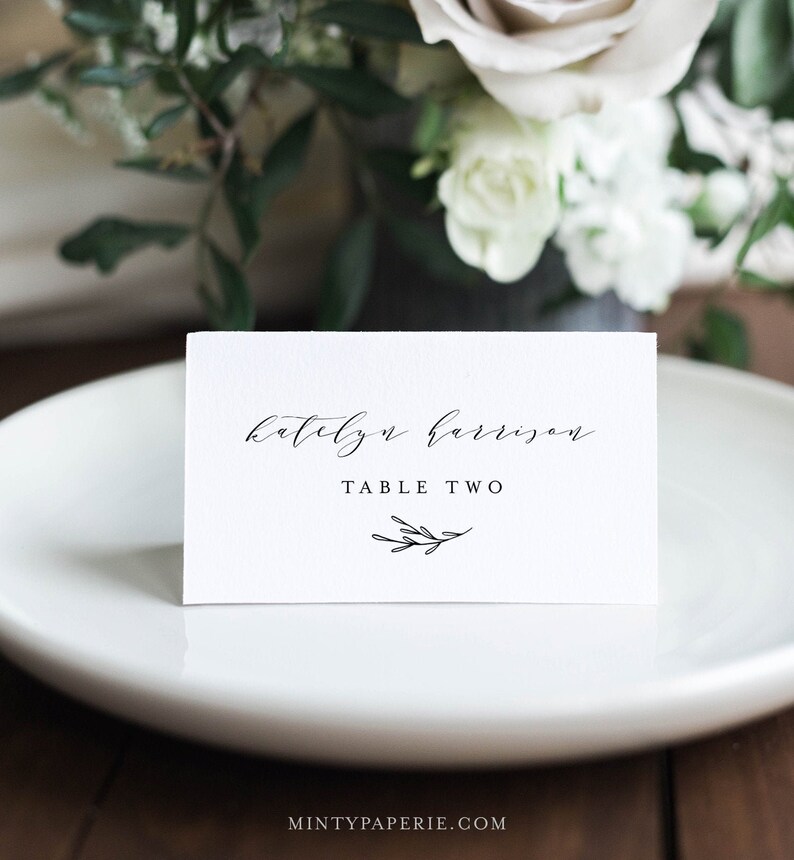 May include: White card with black calligraphy that reads "katelyn harrison" and "TABLE TWO" with a small black twig illustration.