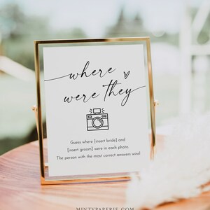 Puede incluir: Un cartel blanco en un marco dorado con el texto "where were they" y un icono de c&aacute;mara. El cartel tambi&eacute;n incluye el texto "Adivina d&oacute;nde [inserta la novia] y [inserta el novio] estaban en cada foto. &iexcl;La persona con la mayor cantidad de respuestas correctas gana!"