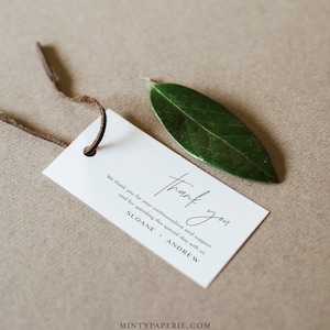 May include: A white thank you tag with black text that reads "We thank you for your continuous love and support and for spending this special day with us. Thank you. Sloane + Andrew." The tag is tied with a brown leather cord and a green leaf is in the background.