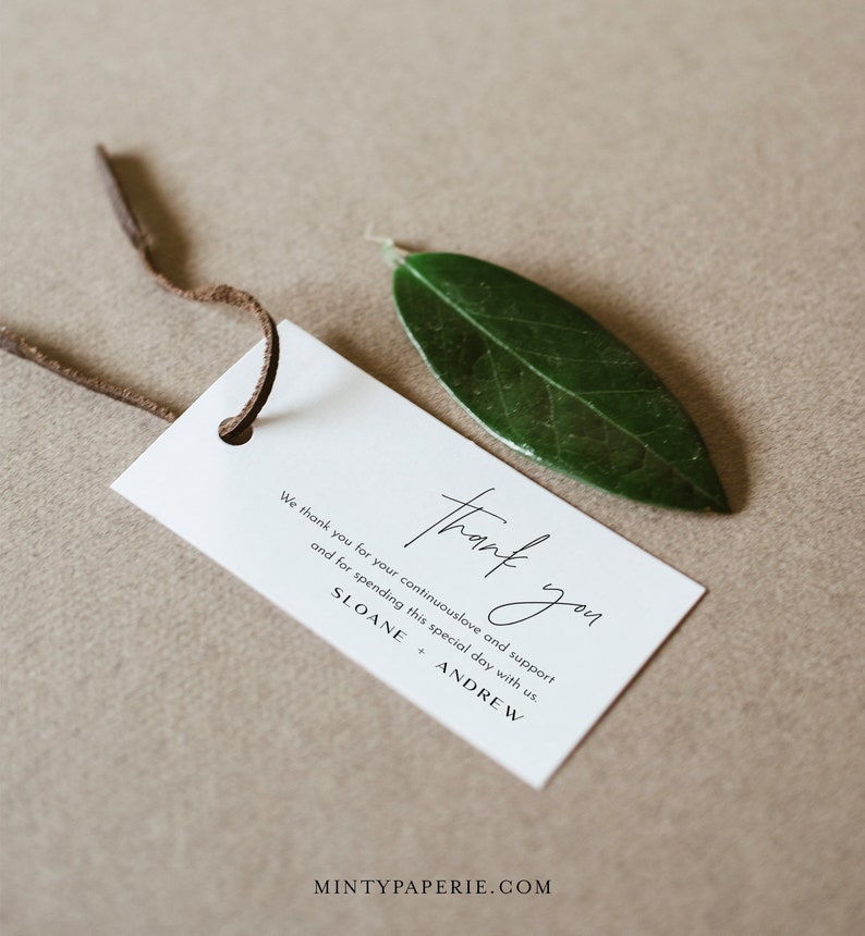 May include: A white thank you tag with black text that reads "We thank you for your continuous love and support and for spending this special day with us. Thank you. Sloane + Andrew." The tag is tied with a brown leather cord and a green leaf is in the background.