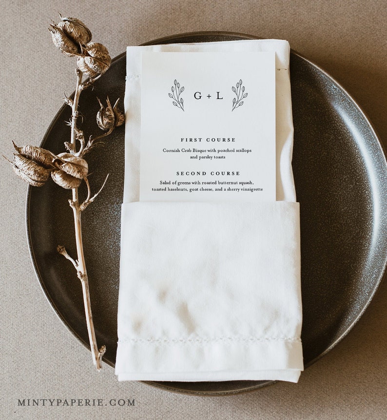 May include: A white folded napkin with a printed menu card on top. The menu card has a simple floral design and the text "G+L" at the top. The menu lists a "First Course" of Cornish Crab Bisque with poached scallops and parsley toasts and a "Second Course" of salad of greens with roasted butternut squash, toasted hazelnuts, goat cheese, and a sherry vinaigrette.
