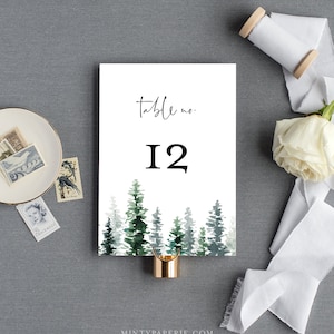May include: White card with black text that reads "table no. 12" and a watercolor illustration of a forest with green trees. The card is held by a gold metal clip.