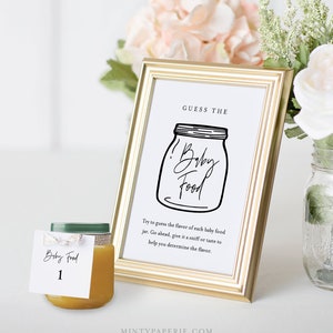 May include: A gold-framed sign with the text "Guess the Baby Food" and a black and white illustration of a jar of baby food. A small glass jar of baby food with a yellow label that says "Baby Food 1" is in the foreground.