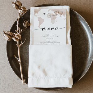 May include: A menu card with a world map design and the word "menu" in cursive. The card is placed in a white napkin on a dark gray plate. The menu lists two courses: Crab Bisque and a salad.