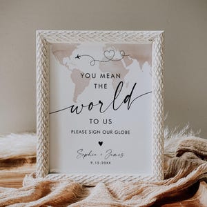 May include: A framed sign with a world map illustration in soft pink tones. The sign reads "YOU MEAN THE WORLD TO US" and includes space for signatures. The frame is white with a textured pattern. The names "Sophia + James" and a date are also included.