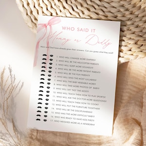 Puede incluir: Una tarjeta de juego para baby shower con el texto "Who Said It?" en escritura rosa, con un lazo rosa. La tarjeta enumera preguntas sobre los padres, con gráficos de bigotes y labios. Las preguntas incluyen quién cambiará los pañales y quién cantará nanas.