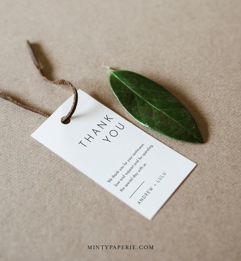 May include: A white thank you tag with black text that reads "THANK YOU" and "We thank you for your continuous love and support and for spending this special day with us." The tag is tied with a brown string and a green leaf is resting on the tag. The text at the bottom of the tag reads "ANDREW + LULU" and "MINTYPAPERIE.COM".