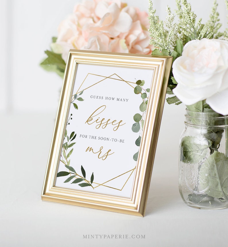 May include: Gold framed sign with a white background and green foliage. The sign says "Guess how many kisses for the soon-to-be Mrs."