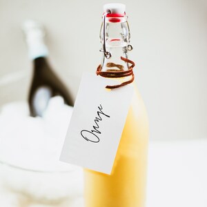May include: A glass bottle with a yellow liquid and a brown leather strap tied around the neck. A white tag with the word "Orange" written in black cursive is attached to the bottle.