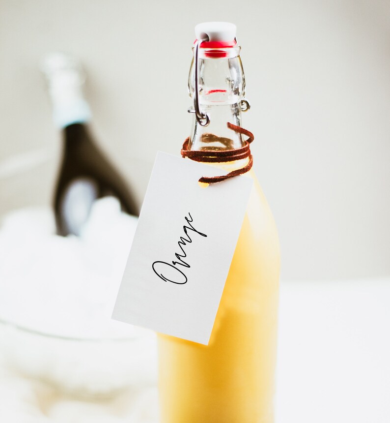 May include: A glass bottle with a yellow liquid and a brown leather strap tied around the neck. A white tag with the word "Orange" written in black cursive is attached to the bottle.