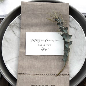 May include: A white card with black calligraphy that reads "Katelyn Harrison" and "Table Two" with a small twig design. The card is placed on a beige linen napkin with a sprig of greenery.