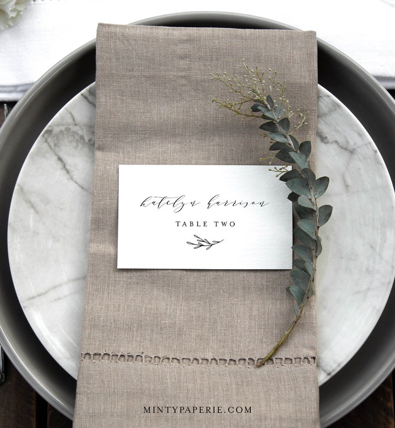 May include: A white card with black calligraphy that reads "Katelyn Harrison" and "Table Two" with a small twig design. The card is placed on a beige linen napkin with a sprig of greenery.