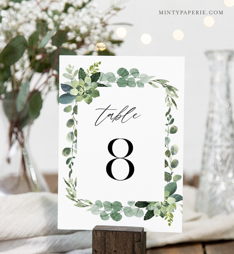 May include: White table number card with a green eucalyptus and succulent wreath border. The card says "Table 8" in black text.