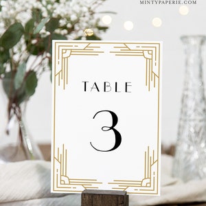 May include: A white table number card with gold art deco style border and accents. The word "TABLE" is printed at the top, with the number "3" in a bold, black font below. The card is displayed in a wooden holder.