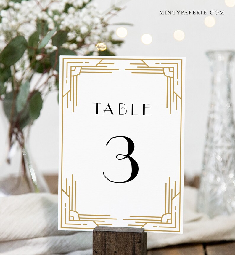 May include: A white table number card with gold art deco style border and accents. The word "TABLE" is printed at the top, with the number "3" in a bold, black font below. The card is displayed in a wooden holder.