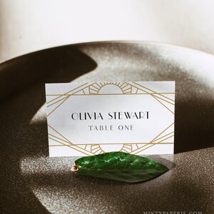 May include: A white place card with gold geometric art deco design, featuring the name "OLIVIA STEWART" and "TABLE ONE". A green leaf rests below the card on a gray surface. The Etsy shop name "MINTYPAPERIE" is in the bottom right corner.