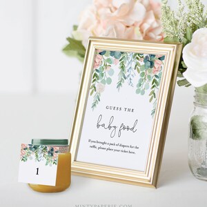 May include: A gold picture frame with a white background and a floral design. The text "GUESS THE baby food" is written in black. The text "If you brought a pack of diapers for the raffle, please place your ticket here." is written in black. A small jar of baby food with a floral design label is in the foreground.