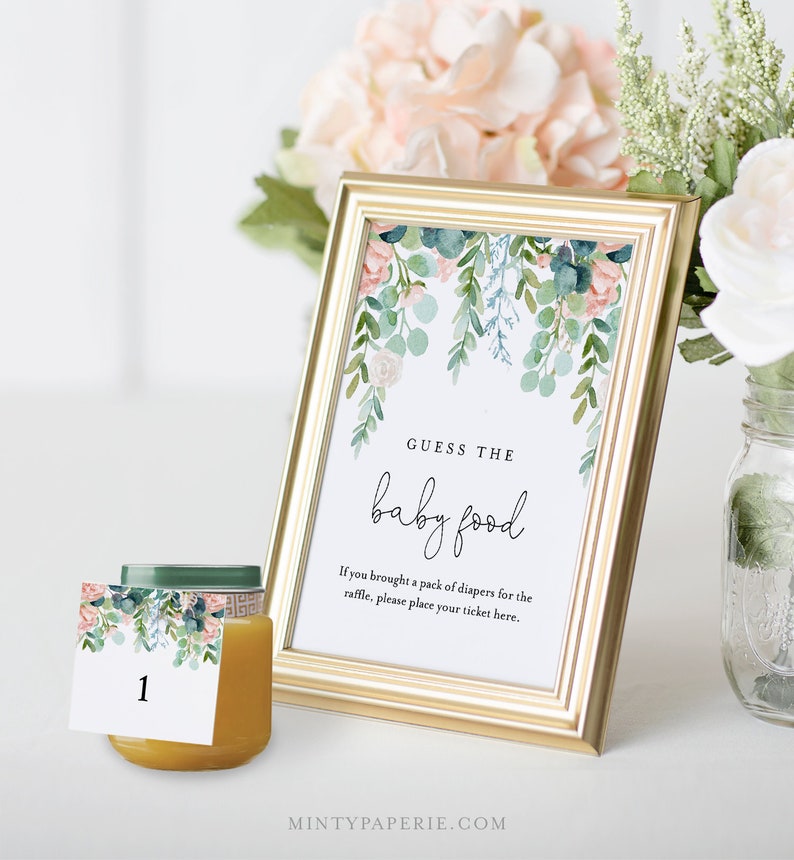 May include: A gold picture frame with a white background and a floral design. The text "GUESS THE baby food" is written in black. The text "If you brought a pack of diapers for the raffle, please place your ticket here." is written in black. A small jar of baby food with a floral design label is in the foreground.