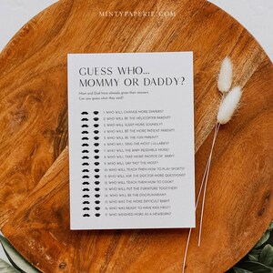 May include: A black and white printable game card with the text "Guess Who... Mommy or Daddy?" and a list of 17 questions about parenting. The questions are printed in black text on a white background. Each question is followed by a row of black lips and mustaches.
