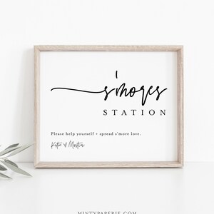 Puede incluir: Un cartel imprimible con el texto "s'mores station" en fuente cursiva negra. El texto "Please help yourself + spread s'more love." está debajo del título. El cartel está enmarcado en un marco de madera claro.