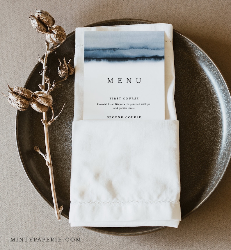 May include: A white napkin with a blue watercolor design menu card tucked inside. The menu card has the word "MENU" in black text and lists "FIRST COURSE" as Cornish Crab Bisque with poached scallops and parsley toasts and "SECOND COURSE" in black text.