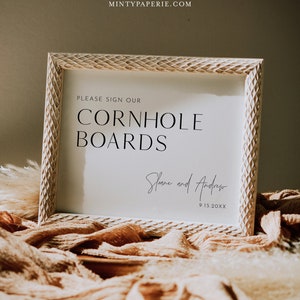 May include: A white sign with a wood-like frame, reading "Please sign our Cornhole Boards" with the names "Sloane and Andrew" and the date "9.15.20XX" in cursive handwriting.