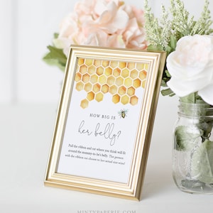 May include: A gold-framed sign with a honeycomb pattern and a bee illustration. The sign reads "HOW BIG IS her belly?" and includes instructions for a baby shower game: "Pull the ribbon and cut where you think will fit around the mommy-to-be's belly. The person with the ribbon cut closest to her actual size wins!"