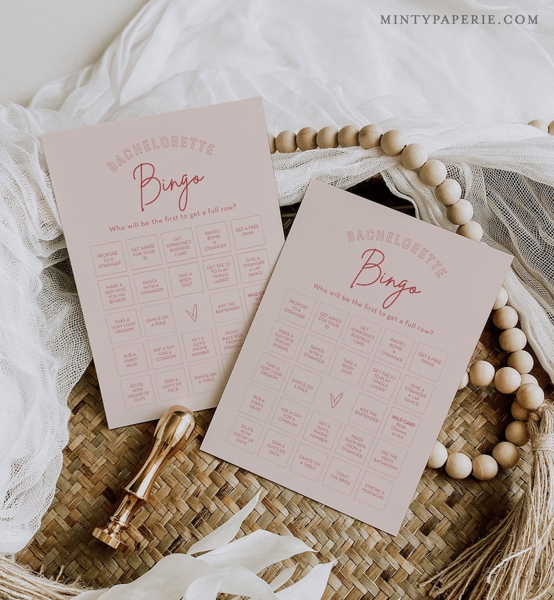 May include: Two pink printable bachelorette bingo cards with a pink heart in the centre. The cards have a list of activities for guests to complete, such as "Get a free drink" and "Dance on a table".