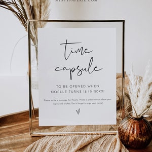 Puede incluir: Un cartel blanco en un marco dorado con el texto "time capsule" y "TO BE OPENED WHEN NOELLE TURNS 18 IN 20XX!"  El cartel también incluye instrucciones para escribir un mensaje para Noelle, hacer una predicción o compartir esperanzas y deseos.  No olvides firmar tu nombre.