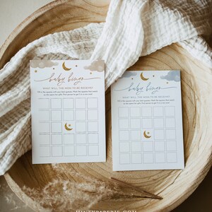 May include: Two blue and gold baby shower bingo cards with a crescent moon and stars design. The text on the cards reads "Baby Bingo" and "What will the mom-to-be receive?"