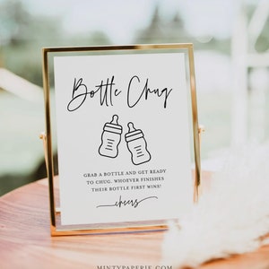 May include: A white sign with a gold frame, announcing a "Bottle Chug" game. The sign features two black and white illustrations of baby bottles and the text "Grab a bottle and get ready to chug. Whoever finishes their bottle first wins! Cheers."
