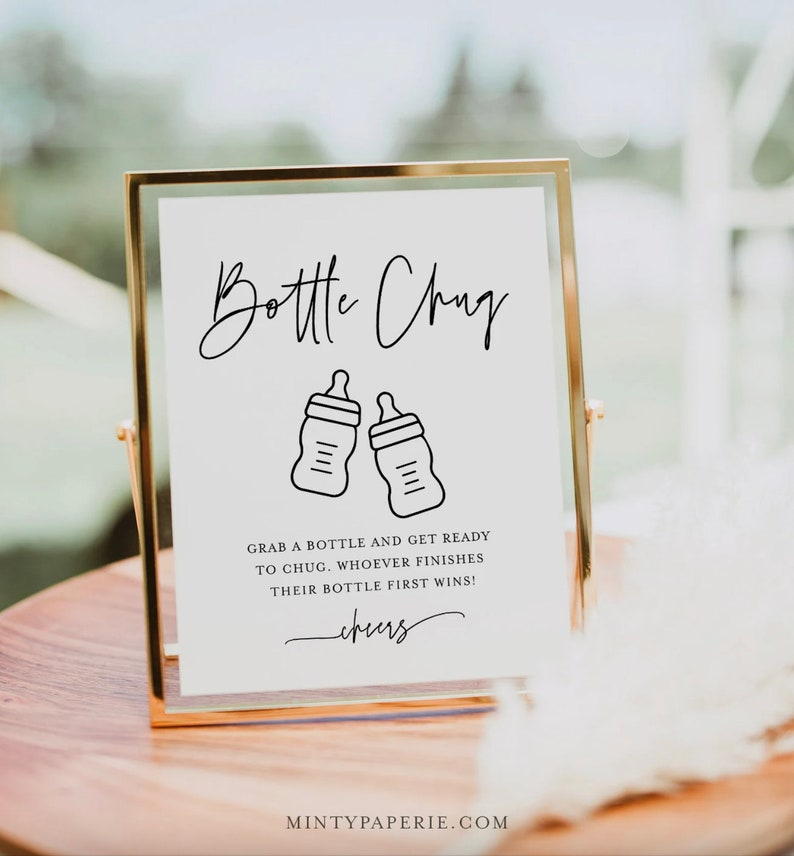 May include: A white sign with a gold frame, announcing a "Bottle Chug" game. The sign features two black and white illustrations of baby bottles and the text "Grab a bottle and get ready to chug. Whoever finishes their bottle first wins! Cheers."