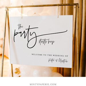 May include: White wedding sign with black calligraphy that reads "the party starts here" and "Welcome to the wedding of Katie & Martin". The sign is hanging from a gold stand.
