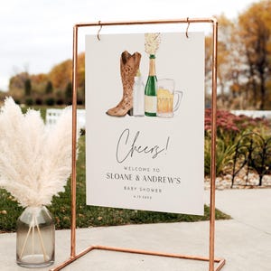 May include: A sign with a watercolor illustration of a cowboy boot, champagne bottle, baby bottle, and beer mug. The sign reads "Cheers!" and welcomes guests to Sloane & Andrew's baby shower.