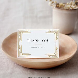 May include: A white thank you card with gold art deco style border and text. The card reads "THANK YOU" above the names "HARPER + ADRIAN". The card is displayed on a wooden bowl.