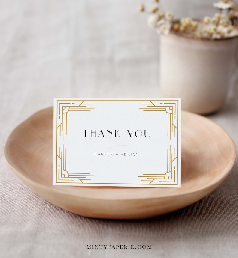 May include: A white thank you card with gold art deco style border and text. The card reads "THANK YOU" above the names "HARPER + ADRIAN". The card is displayed on a wooden bowl.