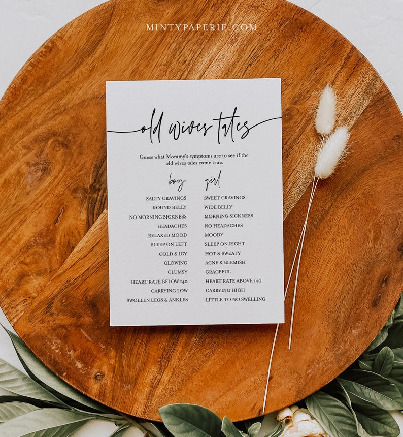 May include: A white card with black text that says "Old Wives Tales" and lists symptoms for a baby boy or girl. The card is on a wooden surface with dried flowers.