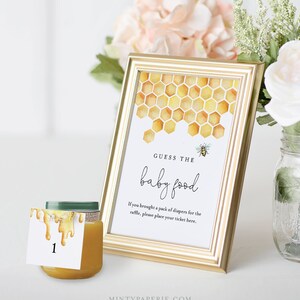May include: A gold frame with a white card inside. The card has a watercolor illustration of a honeycomb with a bee. The text on the card reads "Guess the baby food". There is a small jar of baby food with a number 1 tag on it.