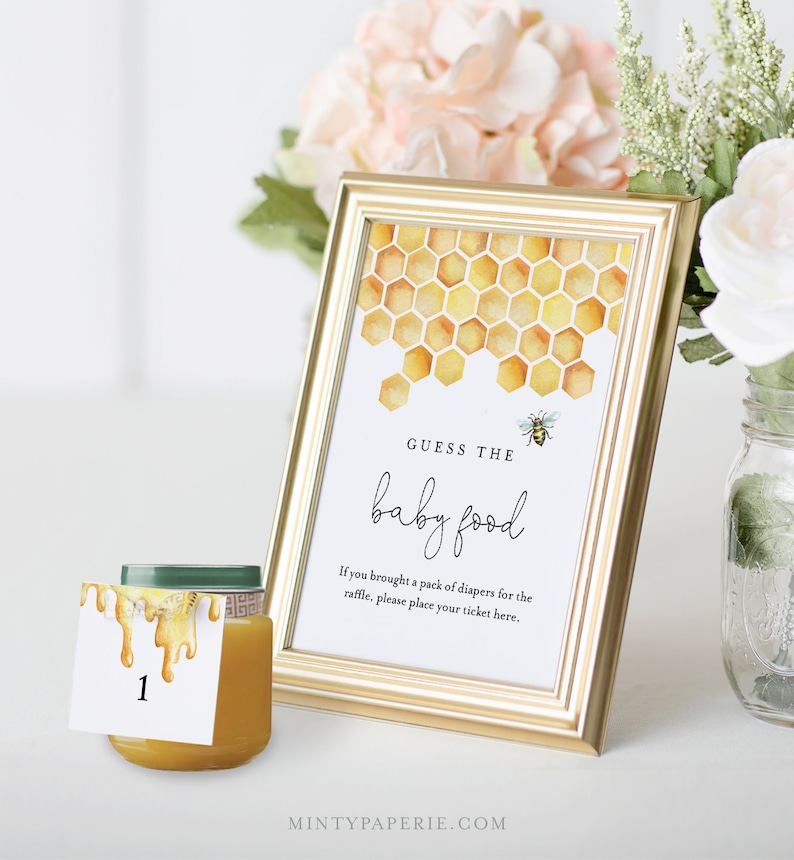 May include: A gold frame with a white card inside. The card has a watercolor illustration of a honeycomb with a bee. The text on the card reads "Guess the baby food". There is a small jar of baby food with a number 1 tag on it.