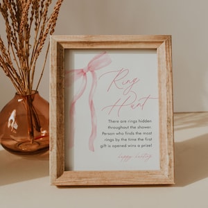 May include: A framed sign with the text "Ring Hunt" in pink script. The sign includes instructions for a ring hunt game, with a pink ribbon design. The frame is made of light-colored wood.