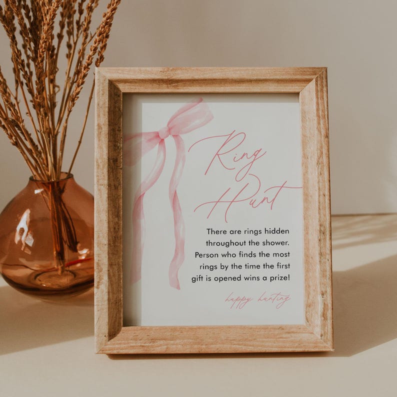 May include: A framed sign with the text "Ring Hunt" in pink script. The sign includes instructions for a ring hunt game, with a pink ribbon design. The frame is made of light-colored wood.